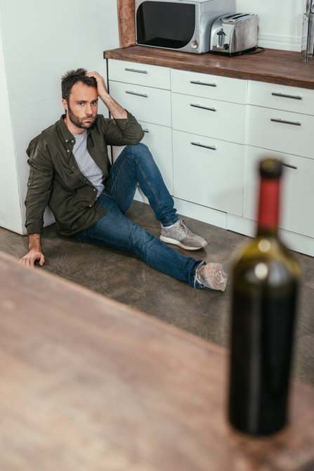 El hombre está sentado en el suelo y mirando la botella de alcohol.